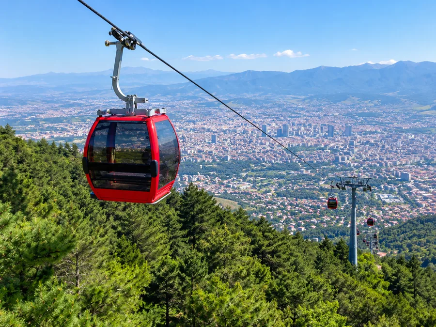 A red Dajti Ekspres gondola rising over pine forest with Tirana in the valley below
